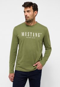 Långärmad skjorta i olivgrön bomull. Har vit text som står "MUSTANG JEANS COMPANY SEDAN 1932" på framsidan. Slät textur.