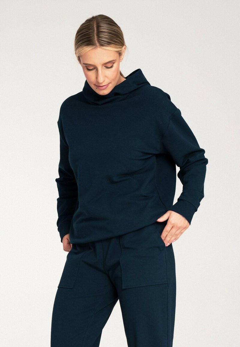 Granatowa bluza z kapturem wykonana z miękkiego materiału, z długimi rękawami i luźnym fasonem. Model łączy ją z pasującymi spodniami, trzymając ręce w kieszeniach.
