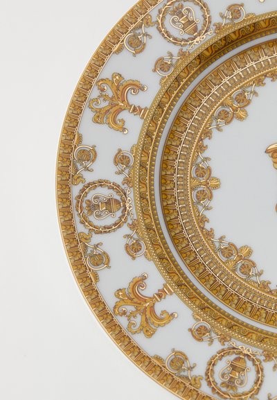 Porcelánový talíř s propracovanými zlatými a hnědými ozdobnými vzory, včetně točítek, květinových motivů a věnců z vavřínu na bílém pozadí.