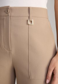 Beige Hosen mit glatter Textur, die über eine Vordertasche, einen Knopfverschluss und ein goldenes quadratisches Schnallendetail verfügen.