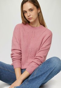 Pull en tricot rose avec un col rond, présentant des motifs texturés et des poignets côtelés, associé à un jean bleu à jambe large.