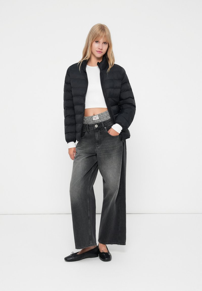 Chaqueta de plumas negra con cremallera y mangas acampanadas, top blanco corto, jeans oscuros de pierna ancha con una cinturilla estampada, planos negros.