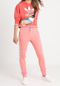 Růžová mikina Adidas s bílým logem, sladěná s odpovídajícími růžovými tepláky se bílými bočními pruhy. Doplněno množstvím barevným ledvinkou.