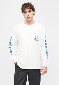 Μακρυμάνικο λευκό t-shirt με τσέπη στο μπροστινό μέρος, με γραφικό σχέδιο, μπλε κείμενο κατά μήκος του μανικιού και χαλαρή εφαρμογή.