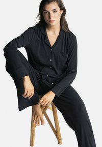 Ensemble de pyjama noir côtelé comprenant un haut à boutons avec une poche et des manches longues, assorti à un pantalon, porté par une personne assise.