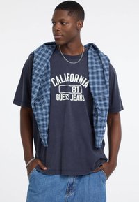 Granatowa koszulka z nadrukiem "California 81 Guess Jeans", w połączeniu z niebieską koszulą w kratę zarzuconą na ramiona, noszona z jasnoniebieskimi szortami dżinsowymi.