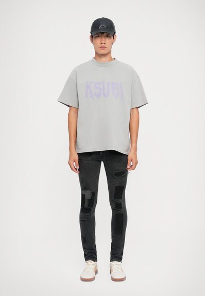 Maglietta grigia oversize con grafica viola "KSUBI", abbinata a jeans skinny neri strappati con toppe e sneakers bianche. Un berretto nero completa il look.