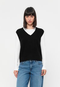 Jonge vrouw met steil donker haar, gekleed in een zwart mouwloos V-halstrui over een witte lange mouwshirt en een spijkerbroek, staand.