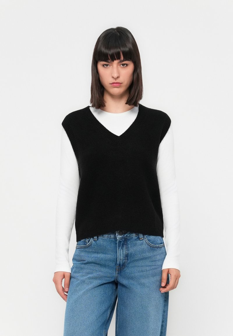Young woman with straight dark hair wearing a black sleeveless V-neck sweater over a white long-sleeve shirt and blue jeans, standing.