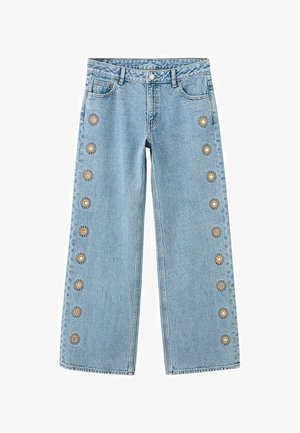 Kobieta w beżowej bluzce bez rękawów i szerokich niebieskich dżinsach z kwiatową haftem, trzymająca brązową torbę na ramię, stoi na szarej podłodze.