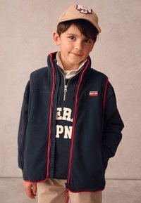 Tamsiai mėlynas vilnonis liemenėlis su raudona apdaila, priekyje su užtrauktuku ir aukštu apykakliu. Dėvimas ant tamsiai mėlyno džemperio su baltais užrašais. Smėlio spalvos kepurė su logotipu.