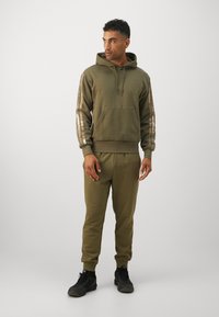 Completo di felpa con cappuccio e pantaloni tuta verde oliva in tessuto morbido, dotato di una tasca anteriore e dettagli a righe sulle maniche. Sneakers nere completano l'outfit.