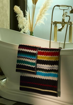 Multicolored towel with a zigzag pattern in shades of red, blue, green, and gray, hanging on a white freestanding bathtub.