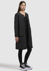 Cappotto lungo trapuntato nero con motivo a diamante, tasche laterali e collo rotondo, abbinato a leggings neri e scarpe da ginnastica alte.