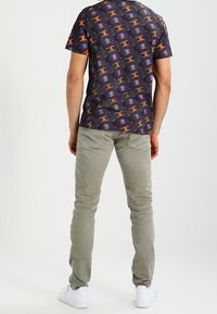 Fioletowa koszulka z krótkim rękawem z wielokolorowym geometrycznym wzorem, zestawiona z dopasowanymi jasnoszarymi spodniami i białymi trampkami.