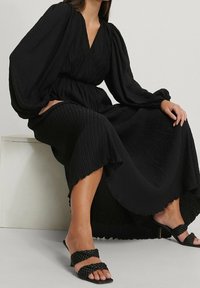 Donna seduta che indossa un lungo vestito nero a trama con maniche a palloncino e sandali con tacco a strappo intrecciato neri.