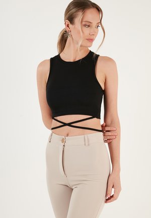 Crni rebrasti crop top s okruglim izrezom i ukriženim naramenicama, uparen s bež uskim hlačama koje imaju zatvaranje na patentni zatvarač i gumb.