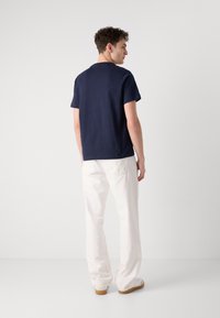 Tricou scurt, de culoare bleumarin, cu guler rotund, asociat cu pantaloni drepți albi și pantofi de culoare deschisă, văzut din spate.