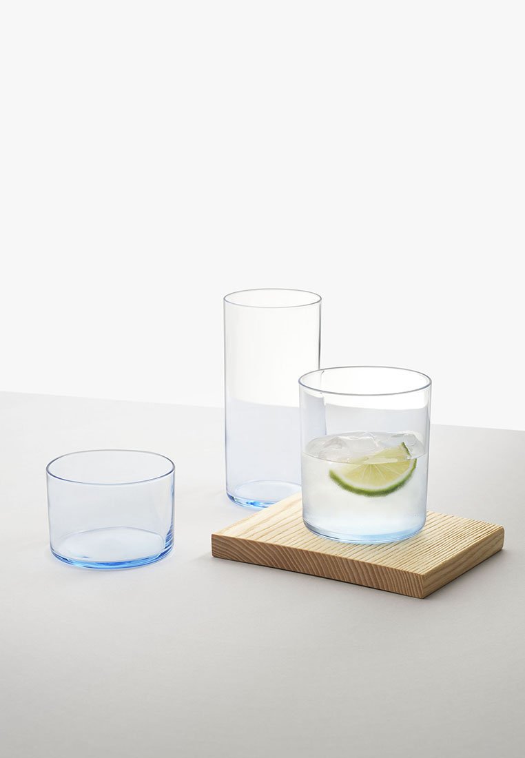 Trzy przezroczyste szklanki z niebieskimi brzegami: jedna krótka z lodem i limonką na drewnianej desce, jedna wysoka pusta i jedna krótka pusta na szarym podłożu.