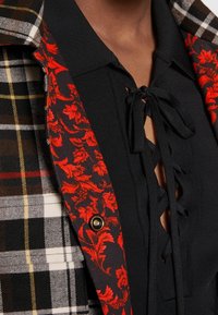 Haut en maille noire avec des liens à lacets devant sous une veste à carreaux doublée d'un tissu rouge à motifs floraux et un bouton doré.