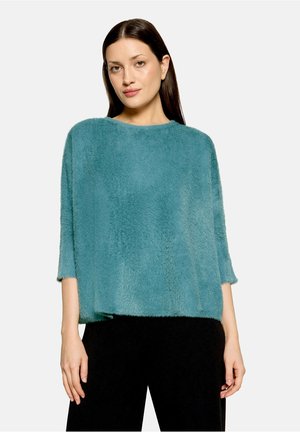Femme aux cheveux raides et foncés portant un pull ample bleu sarcelle duveteux et un pantalon noir, debout devant un fond clair uni.