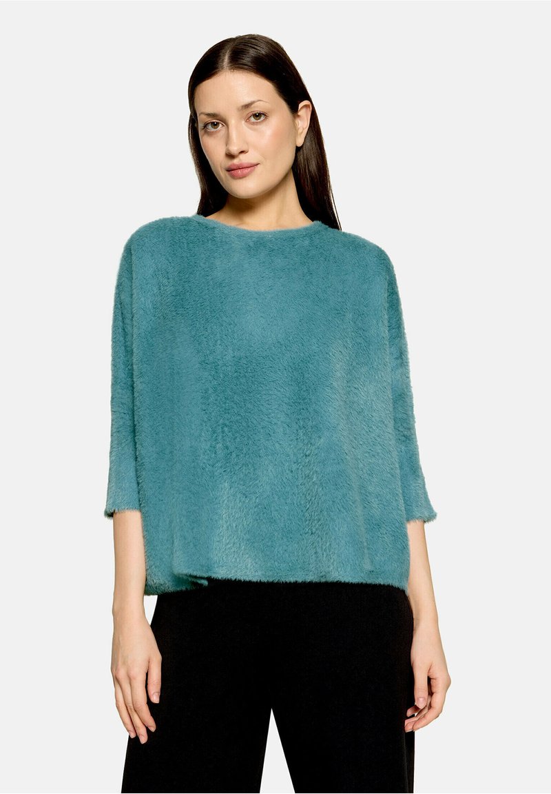 Femme aux cheveux raides et foncés portant un pull ample bleu sarcelle duveteux et un pantalon noir, debout devant un fond clair uni.