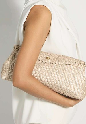 Brazo sosteniendo un bolso de mano tejido beige sobre un fondo de top blanco sin mangas.