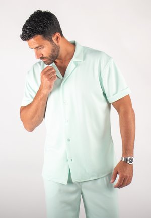 Chemise à manches courtes vert menthe clair avec col, fermeture à boutons et tissu texturé, portée avec un pantalon assorti.