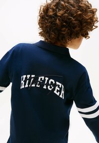 Navy-Polohemd mit weißen gestreiften Ärmeln und dem Text "HILFIGER" in fetter Schrift, ausgestattet mit einem strukturierten gestickten Logo auf der Rückseite.