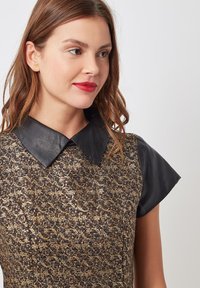 Collar de cuero negro en un vestido de estampado dorado con mangas cortas. La tela texturizada presenta intrincados diseños en espirales.