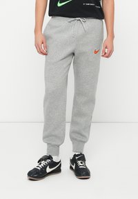 Pelēkie sporta bikses, kas izgatavotas no mīksta kokvilnas auduma, ar elastīgu jostasvietu un auklu, kā arī mazu oranžu Nike logotipu kreisajā augšstilbā.