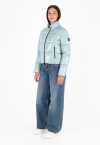 Lichtblauwe gewatteerde jas met een glanzende afwerking, hoge kraag, rits aan de voorkant en zwarte zakritsen, gecombineerd met wijde jeans en witte schoenen.