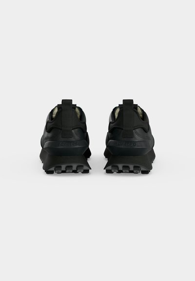 Paire de baskets noires à semelles épaisses, vue arrière, montrant des boucles au talon et des semelles extérieures texturées sur un fond clair uni.