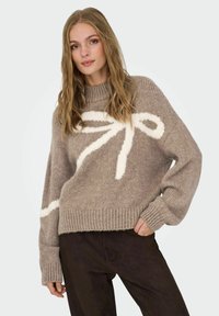 Pull en laine beige tricoté avec un motif de nœud crème. Il présente un col et des poignets côtelés, une coupe ample et une finition texturée.