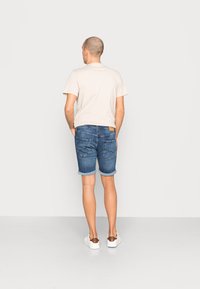 Ljusbeige herr t-shirt ihoplattad med mellanblå denimshorts, med uppvikta ärmar och en läderpatch på baksidan av midjebandet. Vita sneakers.