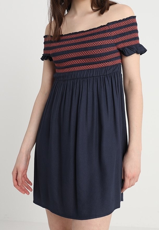 Off-shoulder marinblå klänning med rödrandig smockad livstycke och volangärmar, bärs av en stående person.