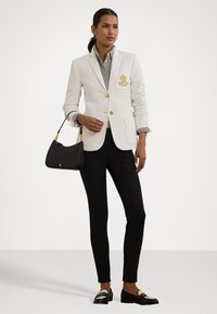 Blazer bianco con bottoni dorati e logo ricamato, indossato sopra una camicia a righe, abbinato a pantaloni neri e mocassini neri. Borsa nera.