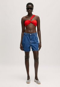 Rode gedraaide bandeau top, hoogzittende blauwe denim shorts en doorschijnende grijze flats. Model draagt zwarte zonnebril en oorbellen met een hoop.