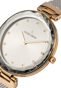 Reloj metálico dorado con bisel de cristal facetado, esfera plateada, marcadores de horas similares a diamantes y correa de malla. Marca: Jacques Lemans.