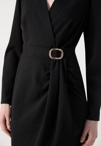 Robe noire avec un décolleté en V profond et des manches longues, présentant un design froncé à la taille maintenu par une boucle ovale métallique. Texture douce.