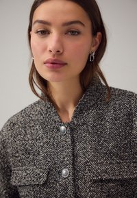 Czarna i biała marynarka w jodełkę z podwyższonym kołnierzem, srebrnymi guzikami i przednimi kieszeniami, wykonana z teksturowanej tkaniny.