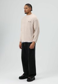 Beige fleece trui met een gestructureerde afwerking, lange mouwen en een zwart geborduurd logo. Gecombineerd met losse zwarte broek en chunky zwarte schoenen.