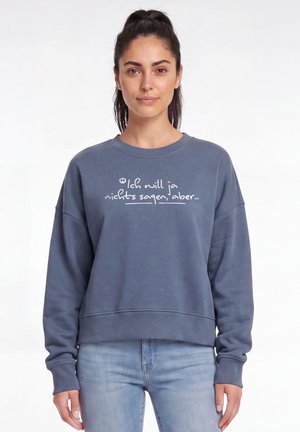 Frau mit dunklem Haar im Pferdeschwanz, die einen blauen Pullover mit deutschem Text "Ich will ja nichts sagen, aber..." und hellblaue Jeans trägt.