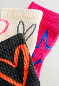 Tre par stickade strumpor: ett svart med ett orange hjärta, ett krämfärgad med ett svart mönster, och ett rosa med en blå zick-zackdesign.