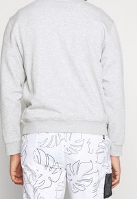 Sudadera gris claro con mangas largas y escote redondo. Material suave, puños y dobladillo acanalados. Combinada con pantalones cortos blancos con patrones de hojas negras.