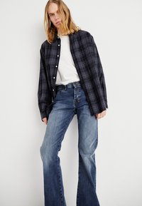 Camisa xadrez preto e cinza sobre uma t-shirt branca, combinada com jeans flare azuis. O conjunto possui botões e um corte descontraído.