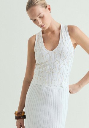 Mujer con top de punto sin mangas blanco con bordado dorado y falda acanalada a juego, posando con la mano en la cadera y mirando hacia abajo.