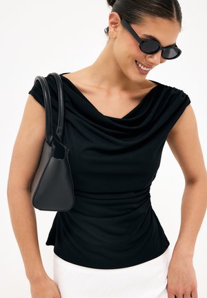 Kvinde iført sort ærmeløs draperet top, sorte solbriller og med en lille sort skuldertaske mod en hvid baggrund.