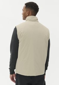 Muškarac s kratkom kovrčavom kosom okrenut sa strane, oblečen u bež prsluk bez rukava preko tamne košulje dugih rukava i crnih hlača na bijeloj pozadini.