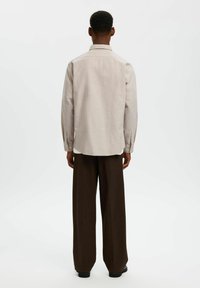 Ανοιχτό μπεζ πουκάμισο με κουμπιά, κλασικό γιακά, μακριά μανίκια και καμπυλωτό τελείωμα, συνδυασμένο με καφέ παντελόνια φαρδιάς γραμμής.